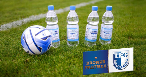 Vier Blankenburger Wiesenquell Mineralwasserflaschen 0,5l stehen auf einem Fußballfeld, daneben liegt ein blau-weißer Fußball. In der rechten unteren Ecke des Bildes: Logo 1. FC Magdeburg Bronzepartner